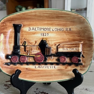 May include: A decorative ceramic plate featuring a vintage train design. The plate is brown with a green border and depicts a black train with red wheels. Text includes "BALTIMORE & OHIO R.R. 1837" and "LAFAYETTE".