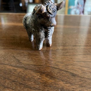 May include: A small, weathered figurine of a bull with horns and a decorative headpiece. The sculpture is primarily gray and brown, with visible texture and detail. The bull stands on four legs, and the overall aesthetic suggests an antique or handcrafted item.