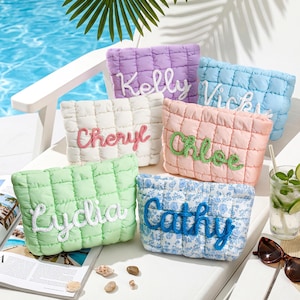 May include: Six quilted cosmetic bags in pastel colors: lavender, white, light blue, peach, green, and blue floral. Each bag features a name embroidered in a contrasting color: Kelly, Cheryl, Vicky, Chloe, Lydia, and Cathy. The bags are displayed on a white chair near a pool.