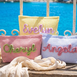 May include: Three rectangular zippered pouches made of seersucker fabric in yellow, red, and blue stripes. Each pouch features a personalized name in raised, textured yarn lettering: Evelyn in lavender, Cheryl in green, and Angela in pink.