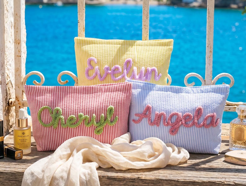 May include: Three rectangular zippered pouches made of seersucker fabric in yellow, red, and blue stripes. Each pouch features a personalized name in raised, textured yarn lettering: Evelyn in lavender, Cheryl in green, and Angela in pink.