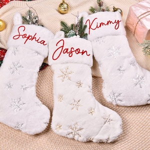 May include: Three white Christmas stockings with snowflake patterns and personalized names in red script: Sophia, Jason, and Kimmy. The stockings are fluffy and decorated with silver and gold snowflakes, perfect for holiday decor.
