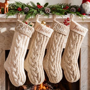 May include: Four cream-colored cable knit Christmas stockings hanging in a row. Each stocking features a ribbed cuff with a name embroidered in cursive script: Elera, Maisie, Robin, and Jason. The stockings are made of a thick, textured yarn.