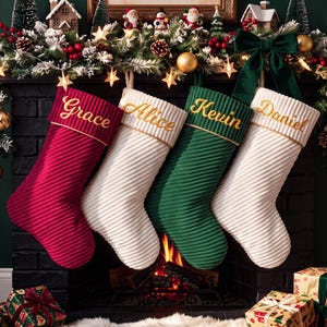 Peut inclure: Quatre chaussettes de Noël personnalisées en tissu côtelé rouge foncé, crème, vert foncé et crème. Chaque chaussette présente un revers avec les prénoms Grace, Alice, Kevin et Daniel brodés en fil doré. Les chaussettes sont suspendues.