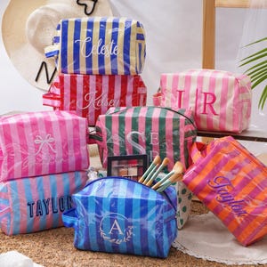 May include: A collection of rectangular cosmetic bags featuring vertical stripes in various colours including blue, yellow, red, pink, green, and orange. Each bag is personalised with embroidered names or initials in white or dark thread.