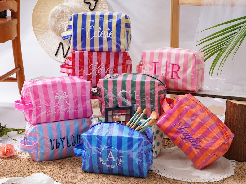 May include: A collection of rectangular cosmetic bags featuring vertical stripes in various colours including blue, yellow, red, pink, green, and orange. Each bag is personalised with embroidered names or initials in white or dark thread.