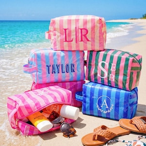 May include: A collection of rectangular toiletry bags with striped patterns in pink, blue, and green. The bags feature personalised text including LR, TAYLOR, SEB, and A. One open bag contains sunscreen bottles and sunglasses, with sandals nearby.