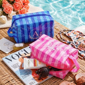 May include: Two rectangular cosmetic bags with a glossy finish and vertical stripes. One bag is blue with a circular monogram featuring the letter A, and the other is pink with a white embroidered bow design. Both bags have a side carry handle.