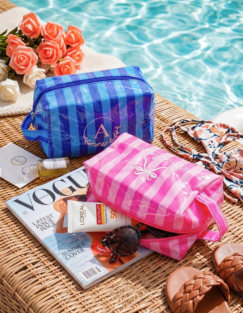 May include: Two rectangular cosmetic bags with a glossy finish and vertical stripes. One bag is blue with a circular monogram featuring the letter A, and the other is pink with a white embroidered bow design. Both bags have a side carry handle.
