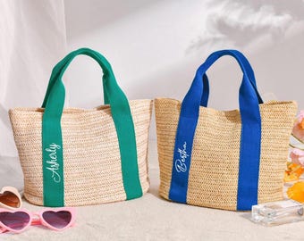 May include: Two woven tote bags with contrasting handles. One has green handles and the name "Asherly" in white script. The other has blue handles with the name "Bertha". Both bags are beige and sit on a light surface.