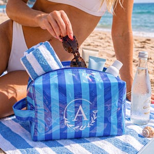 May include: A blue and light blue striped cosmetic bag with a side handle and a white circular monogram featuring the letter A surrounded by a leaf wreath. The bag is filled with a white and blue striped towel, sunglasses, and various skincare tubes.