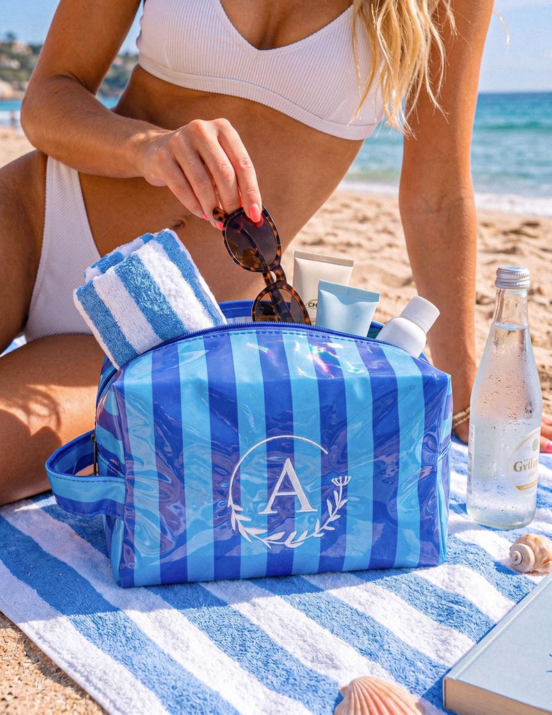 May include: A blue and light blue striped cosmetic bag with a side handle and a white circular monogram featuring the letter A surrounded by a leaf wreath. The bag is filled with a white and blue striped towel, sunglasses, and various skincare tubes.