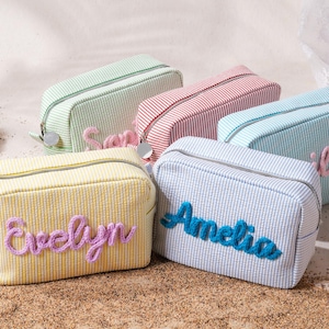 May include: A collection of five rectangular zippered pouches made of striped fabric in pastel yellow, light green, red, and blue. Each pouch features a personalized name embroidered in raised, textured yarn lettering on the front panel.