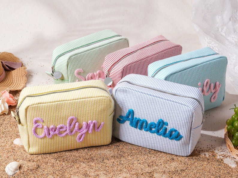 May include: A collection of five rectangular zippered pouches made of striped fabric in pastel yellow, light green, red, and blue. Each pouch features a personalized name embroidered in raised, textured yarn lettering on the front panel.