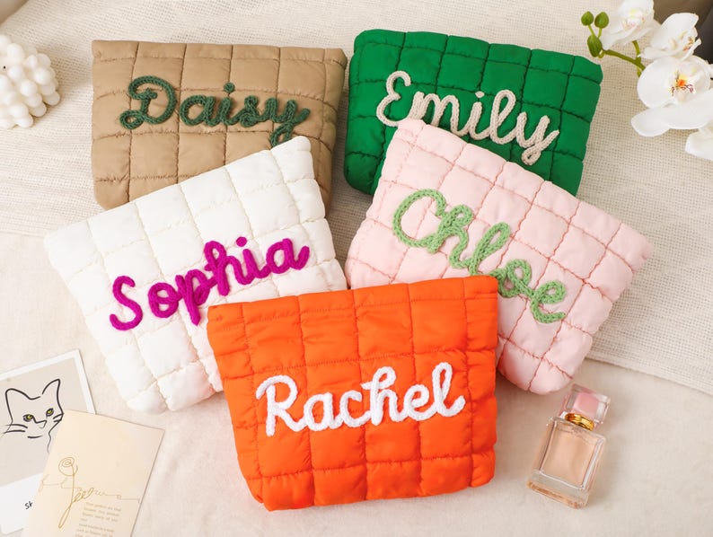May include: Five quilted pouches in various colors: tan, green, white, pink, and orange. Each pouch features a different name embroidered in a contrasting color: Daisy, Emily, Sophia, Chloe, and Rachel. The pouches are arranged on a light-colored surface.