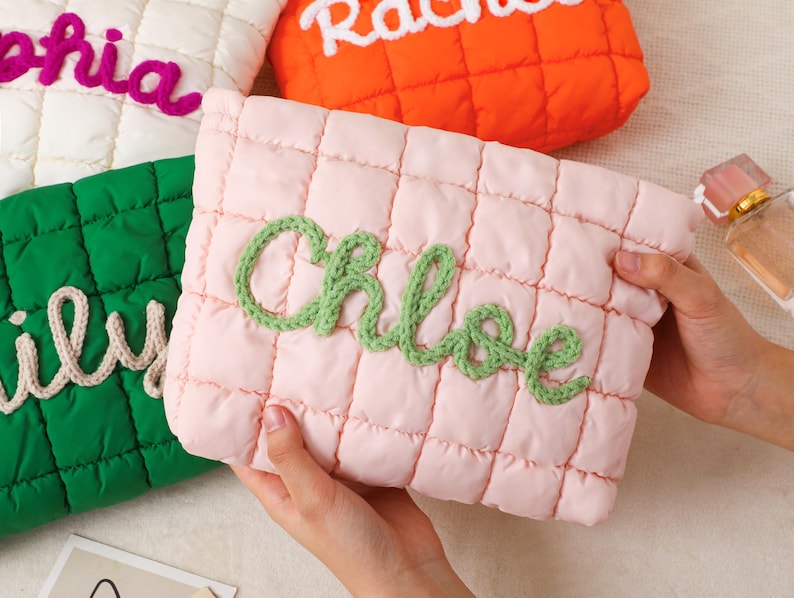 May include: A collection of quilted cosmetic bags in various colors, each embroidered with a name. The bags are in white, orange, green, and pink. The pink bag has the name "Chloe" in green yarn. The bags are rectangular with a quilted pattern.