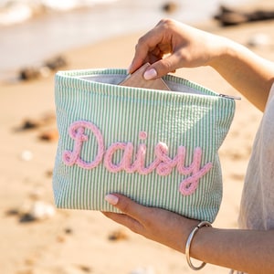 May include: A rectangular zippered pouch featuring a light green and white seersucker striped pattern. The front is decorated with the name Daisy written in thick, textured, light pink yarn lettering. The pouch has a silver zipper closure.