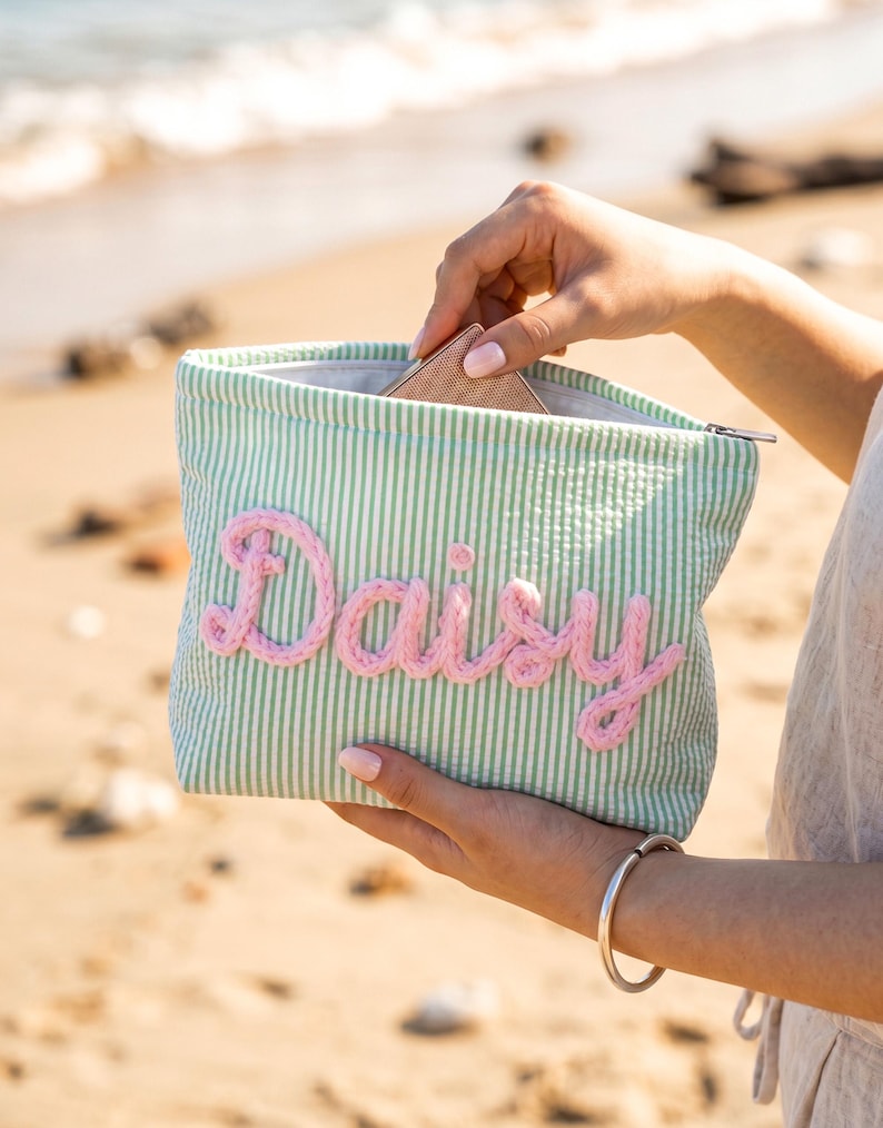 May include: A rectangular zippered pouch featuring a light green and white seersucker striped pattern. The front is decorated with the name Daisy written in thick, textured, light pink yarn lettering. The pouch has a silver zipper closure.