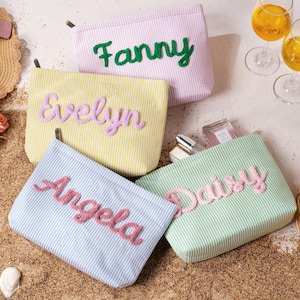 May include: Four rectangular fabric pouches with white pinstripes in lavender, yellow, light blue, and light green. Each pouch features a name embroidered in a raised, textured script: Fanny in green, Evelyn in purple, Angela in pink, and Daisy in pink.