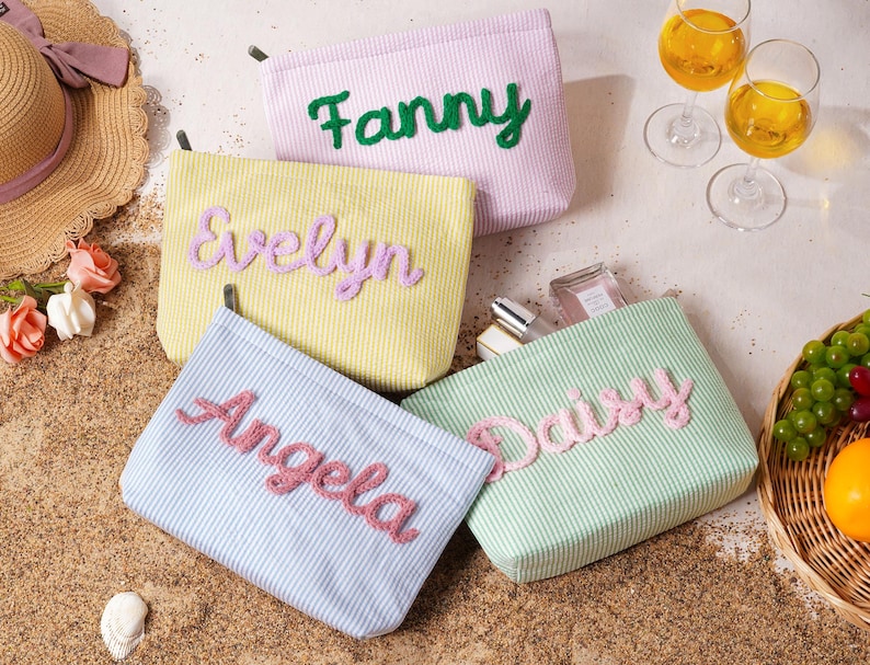 May include: Four rectangular fabric pouches with white pinstripes in lavender, yellow, light blue, and light green. Each pouch features a name embroidered in a raised, textured script: Fanny in green, Evelyn in purple, Angela in pink, and Daisy in pink.