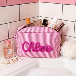 May include: A pink and white striped fabric toiletry bag with the name Chloe embroidered in bright pink yarn on the front. The bag is filled with various cosmetic bottles and containers, including a perfume bottle labeled COOC Chanel.