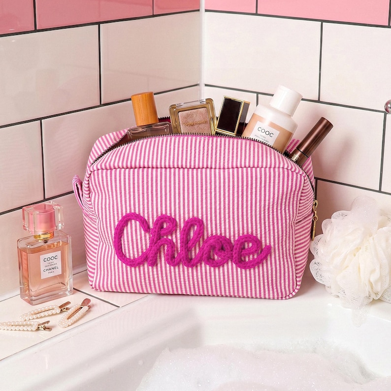 May include: A pink and white striped fabric toiletry bag with the name Chloe embroidered in bright pink yarn on the front. The bag is filled with various cosmetic bottles and containers, including a perfume bottle labeled COOC Chanel.