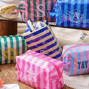 May include: A collection of rectangular, zipped cosmetic bags made of glossy, striped material in various colour combinations including pink, blue, green, and yellow, each featuring personalised embroidered names or initials on the front.