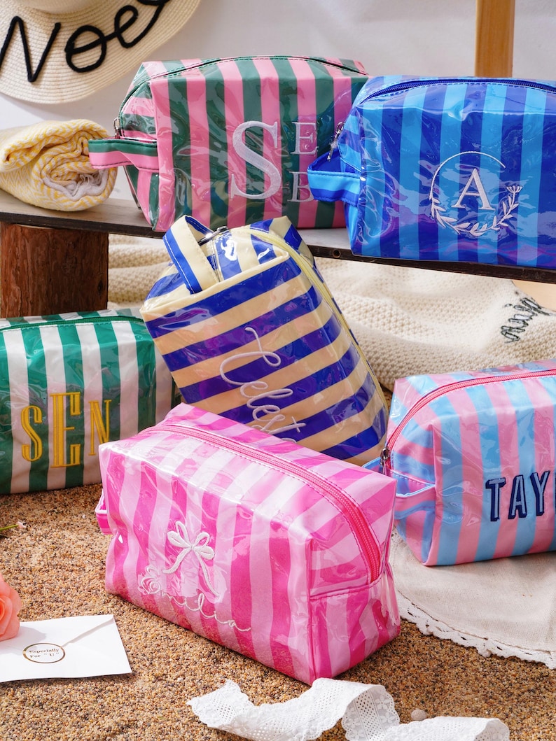 May include: A collection of rectangular, zipped cosmetic bags made of glossy, striped material in various colour combinations including pink, blue, green, and yellow, each featuring personalised embroidered names or initials on the front.