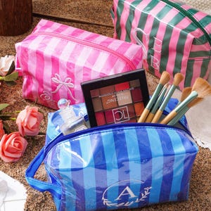 May include: Three rectangular cosmetic bags with striped patterns in pink, green, and blue. The blue bag features a white letter A monogram, the pink bag has a white bow embroidery, and the green bag displays white SB initials. Makeup brushes and a palette are inside.