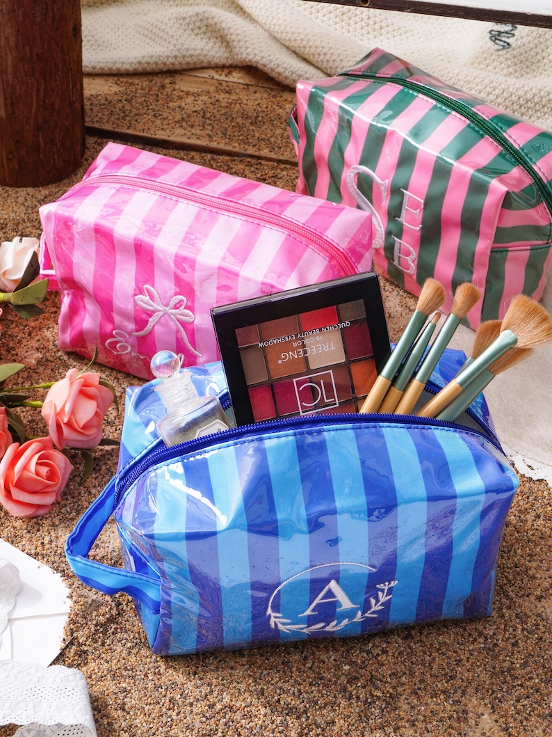 May include: Three rectangular cosmetic bags with striped patterns in pink, green, and blue. The blue bag features a white letter A monogram, the pink bag has a white bow embroidery, and the green bag displays white SB initials. Makeup brushes and a palette are inside.
