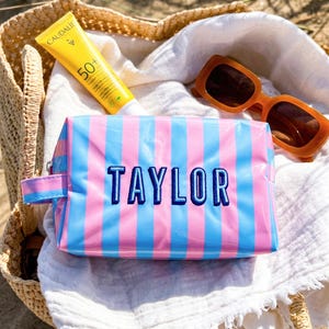 May include: A rectangular cosmetic bag with a glossy finish featuring vertical pink and blue stripes. The name TAYLOR is embroidered in dark blue thread on the front. A small side handle is attached to the left side of the zippered pouch.