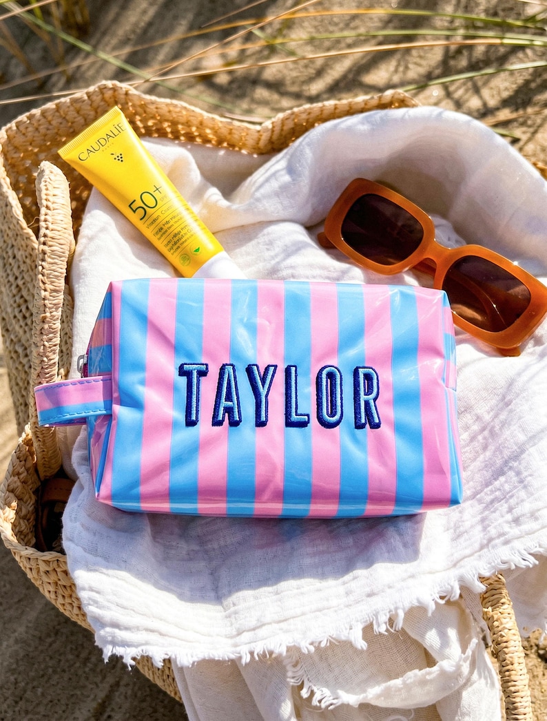 May include: A rectangular cosmetic bag with a glossy finish featuring vertical pink and blue stripes. The name TAYLOR is embroidered in dark blue thread on the front. A small side handle is attached to the left side of the zippered pouch.