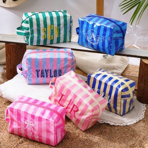 May include: Six rectangular cosmetic bags with vertical stripes in various colours including green, blue, pink, and yellow. Each bag features a zippered closure, a side handle, and personalised embroidered text or monograms on the front surface.