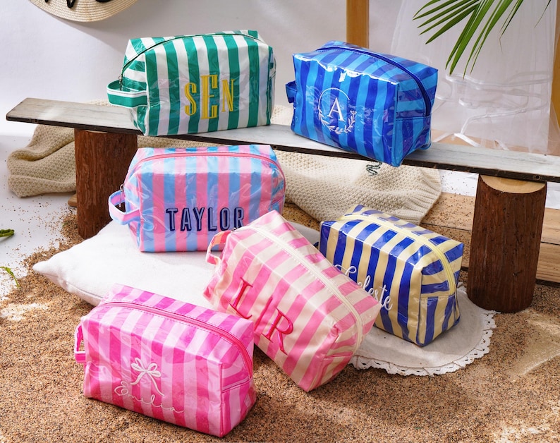 May include: Six rectangular cosmetic bags with vertical stripes in various colours including green, blue, pink, and yellow. Each bag features a zippered closure, a side handle, and personalised embroidered text or monograms on the front surface.