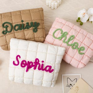 May include: Three quilted cosmetic bags in tan, pink, and white. Each bag has a name embroidered in a contrasting color: "Daisy" in green, "Chloe" in green, and "Sophia" in fuchsia. The bags are rectangular with a quilted pattern.