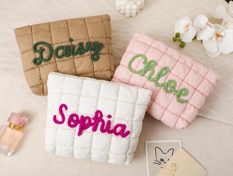 May include: Three quilted cosmetic bags in tan, pink, and white. Each bag has a name embroidered in a contrasting color: "Daisy" in green, "Chloe" in green, and "Sophia" in fuchsia. The bags are rectangular with a quilted pattern.