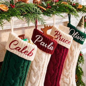 May include: Four knit Christmas stockings hanging in a row, featuring cable-knit patterns and white cuffs embroidered with the names Caleb, Paula, Bruce, and Olivia. The stockings are green, cream, and red, with a wooden reindeer ornament.