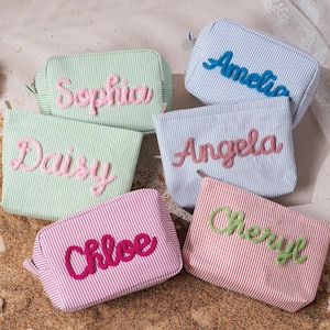May include: A collection of six rectangular zippered pouches made of striped fabric in pastel green, light blue, and pink. Each pouch features a name embroidered in a raised, textured script: Sophia, Amelia, Daisy, Angela, Chloe, and Cheryl.