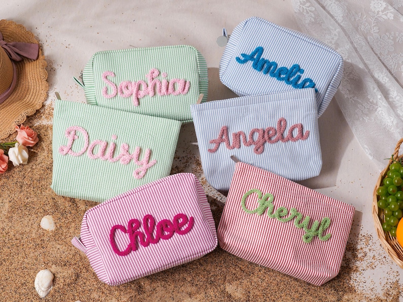 May include: A collection of six rectangular zippered pouches made of striped fabric in pastel green, light blue, and pink. Each pouch features a name embroidered in a raised, textured script: Sophia, Amelia, Daisy, Angela, Chloe, and Cheryl.