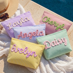 May include: Five rectangular seersucker pouches in yellow, light blue, lavender, mint green, and pink, each featuring a name embroidered in raised, fuzzy script lettering. The names are Evelyn, Angela, Layla, Daisy, and Cheryl, displayed on striped fabric.