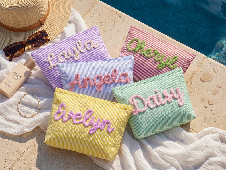 May include: Five rectangular seersucker pouches in yellow, light blue, lavender, mint green, and pink, each featuring a name embroidered in raised, fuzzy script lettering. The names are Evelyn, Angela, Layla, Daisy, and Cheryl, displayed on striped fabric.