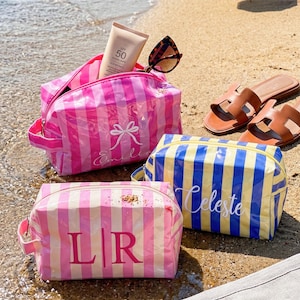 May include: Three rectangular cosmetic bags with striped patterns. One bag is pink and white with a white bow and the name Emilia. Another is yellow and blue with the name Celeste. The third is pink and white with the monogram L|R.