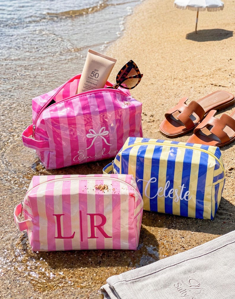 May include: Three rectangular cosmetic bags with striped patterns. One bag is pink and white with a white bow and the name Emilia. Another is yellow and blue with the name Celeste. The third is pink and white with the monogram L|R.
