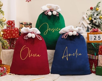 May include: Three velvet Christmas gift bags in red, green, and blue, each with a white faux fur trim and a drawstring closure. The bags are personalized with gold script names: Olivia, Jessica, and Amelia. Festive holiday decor surrounds the bags.