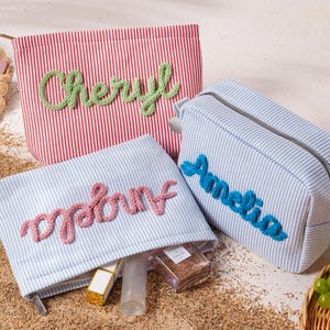 May include: Three rectangular zippered pouches made of striped fabric. One red and white striped pouch features the name Cheryl in green yarn. Two light blue and white striped pouches feature the names Angela and Amelia in pink and blue yarn.