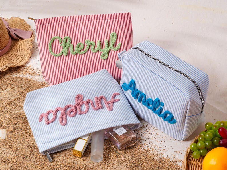 May include: Three rectangular zippered pouches made of striped fabric. One red and white striped pouch features the name Cheryl in green yarn. Two light blue and white striped pouches feature the names Angela and Amelia in pink and blue yarn.