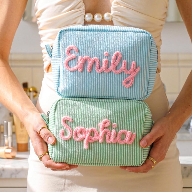May include: Two rectangular zippered pouches made of seersucker fabric. The top pouch is light blue and white striped with the name Emily in pink yarn lettering. The bottom pouch is light green and white striped with the name Sophia in pink yarn.