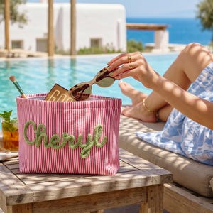 May include: A rectangular zippered pouch featuring a pink and white vertical striped pattern with the name Cheryl embroidered in green cursive script on the front. The pouch contains a makeup brush, a small brown palette, and tortoise shell sunglasses.