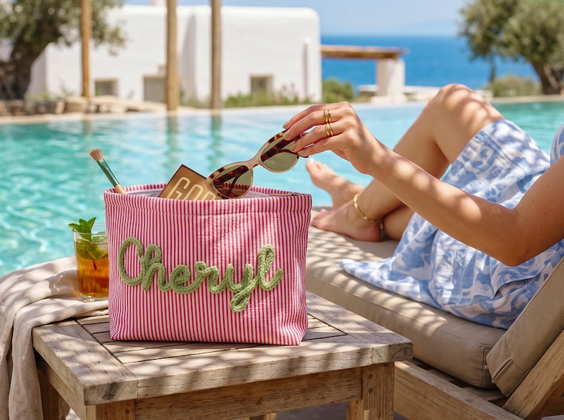 May include: A rectangular zippered pouch featuring a pink and white vertical striped pattern with the name Cheryl embroidered in green cursive script on the front. The pouch contains a makeup brush, a small brown palette, and tortoise shell sunglasses.
