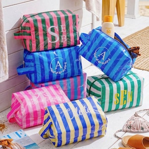 May include: A collection of rectangular, striped toiletry bags in various colour combinations including pink and green, blue and light blue, and yellow and blue. Each bag features a zippered closure, a side handle, and personalised lettering.